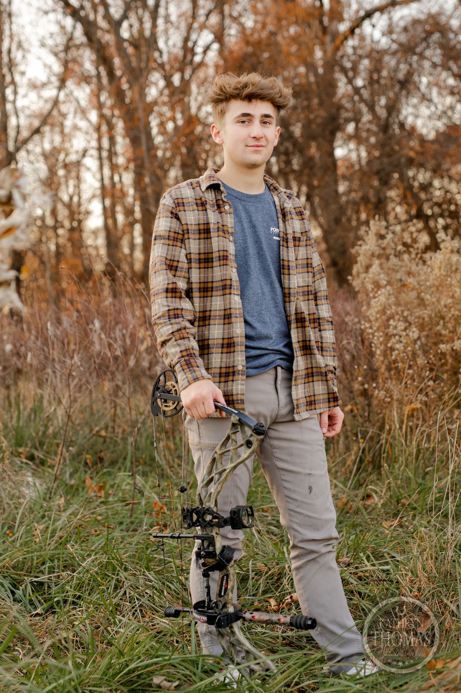 A person in a plaid shirt poses with a compound bow in a field during autumn.