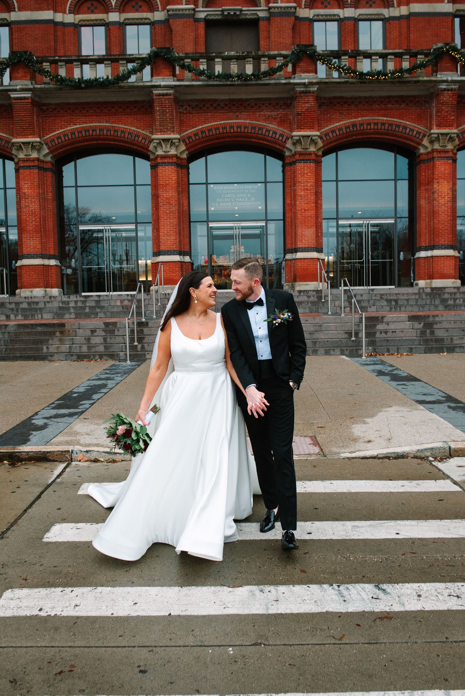 Elegant wedding couple poses in front of historic red brick building with arched windows.