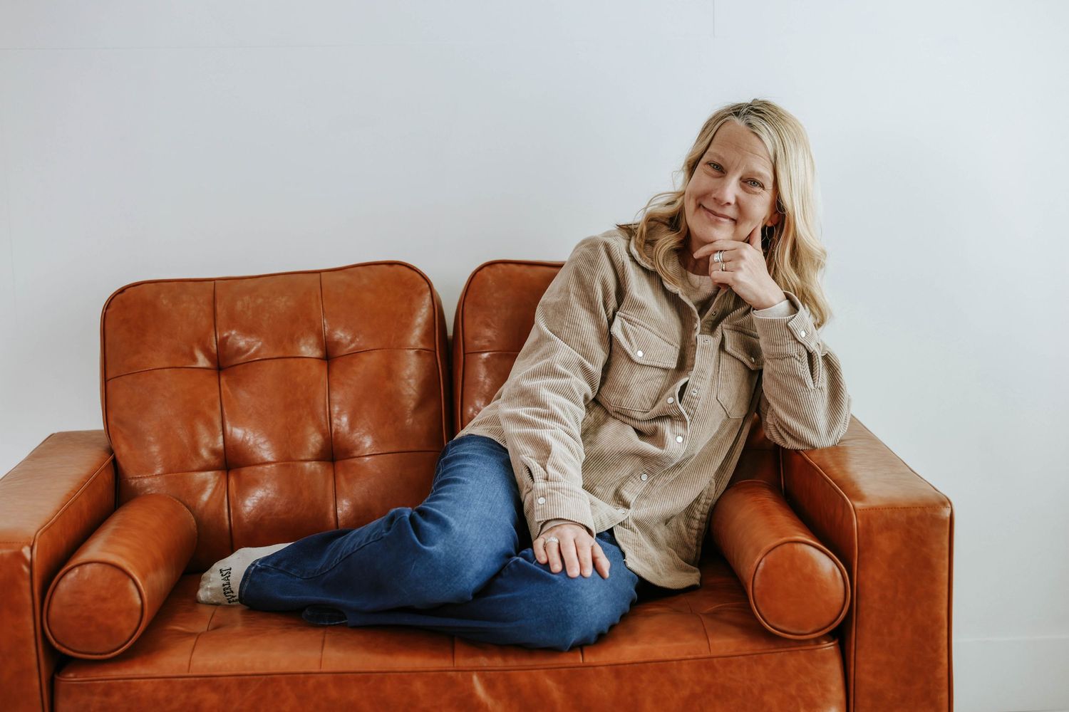 Person in beige jacket and jeans relaxes on vintage brown leather couch against white wall.