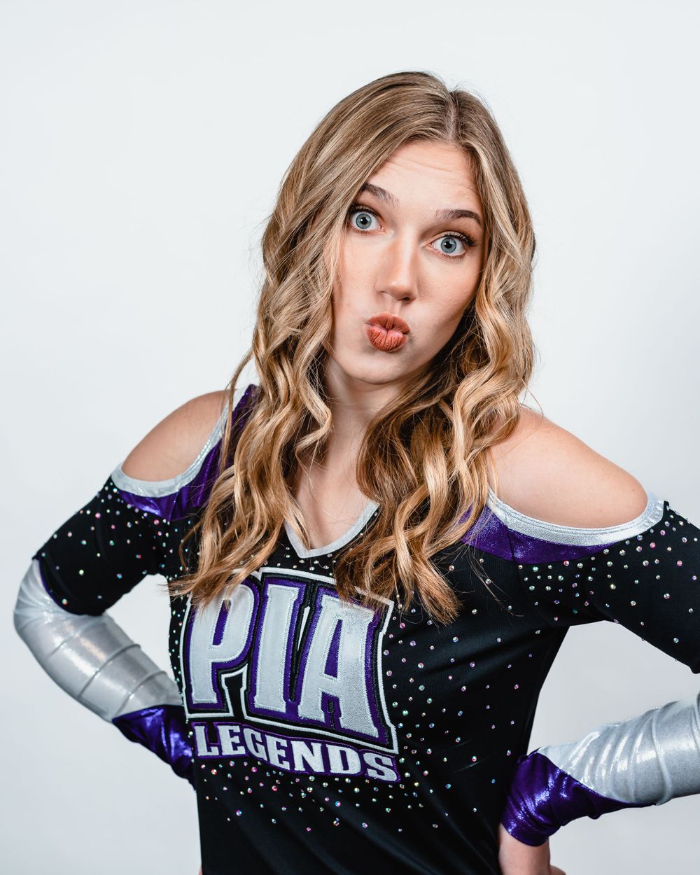 Cheerleader in sparkly purple uniform strikes playful pose against white background.