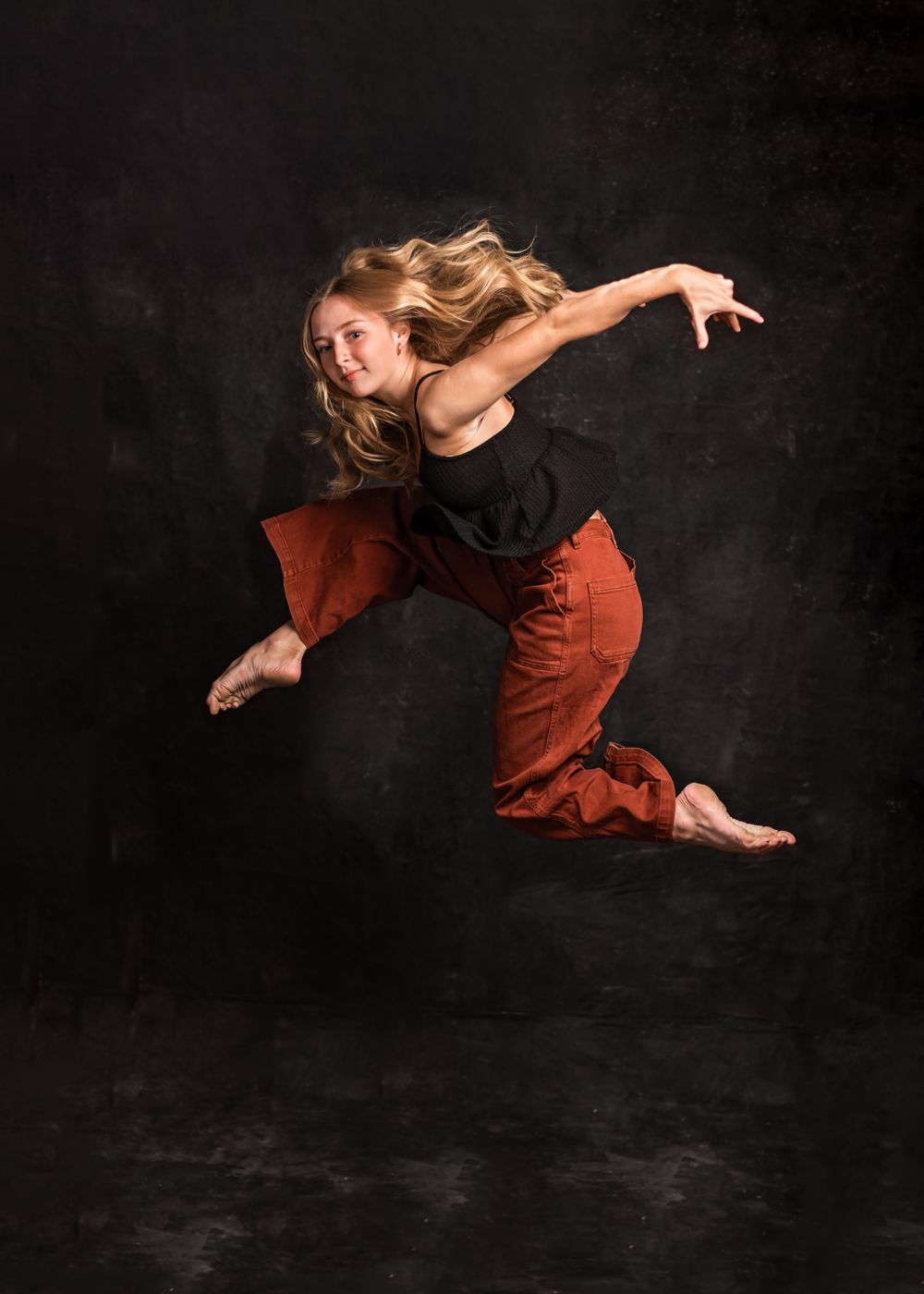 Dancer in black top and rust pants leaps gracefully against dark background.