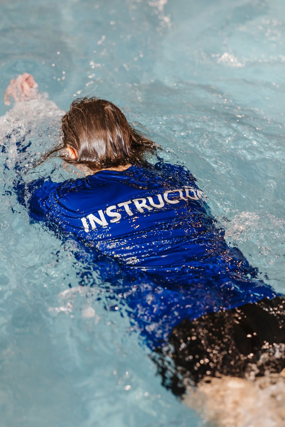 Swimming instructor in blue uniform demonstrates proper backstroke technique in a clear pool during training session.