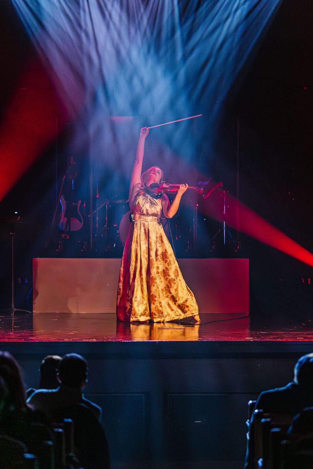 A silhouetted audience watches a performer in a golden dress on stage illuminated by dramatic blue and red spotlights.