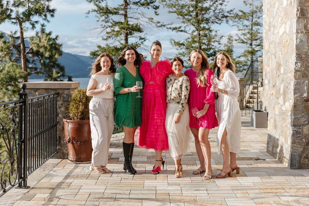 A group of women pose together on a scenic terrace wearing colorful dresses in pink, green and white.