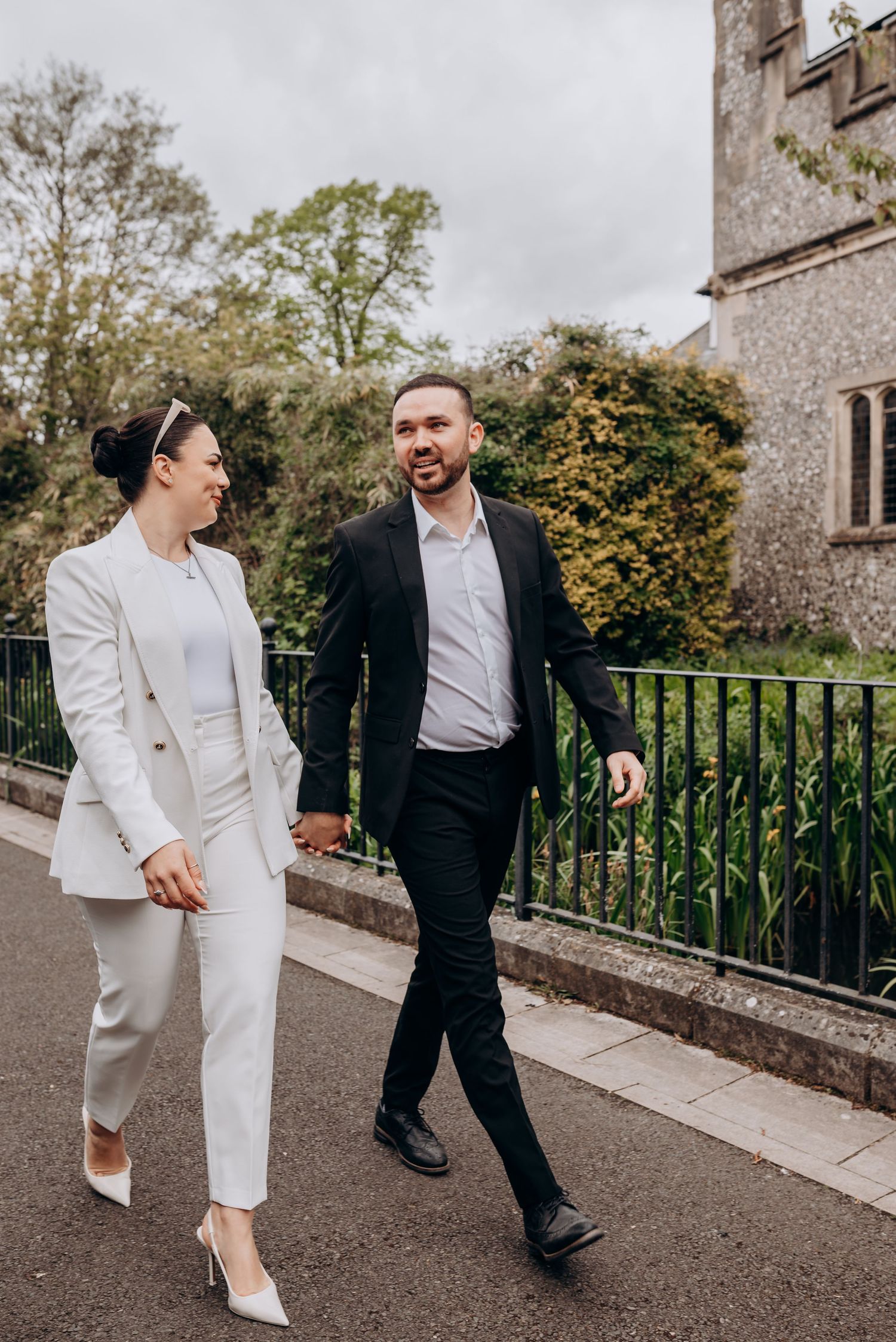 A couple walks together on a path outside a stone building, wearing contrasting black and white formal attire.