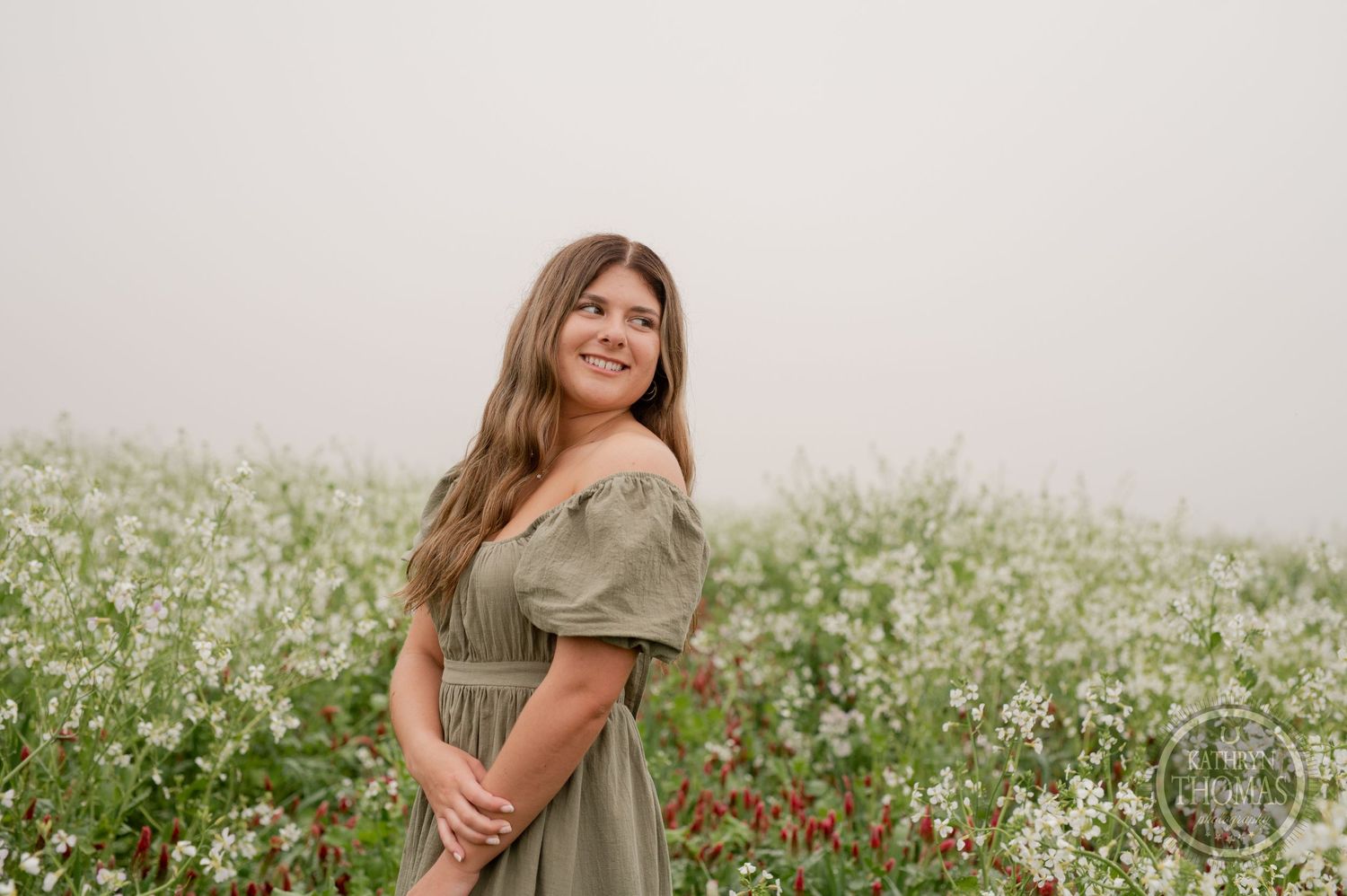 Person in sage green off-shoulder dress poses in a meadow of white wildflowers during a dreamy outdoor photoshoot.