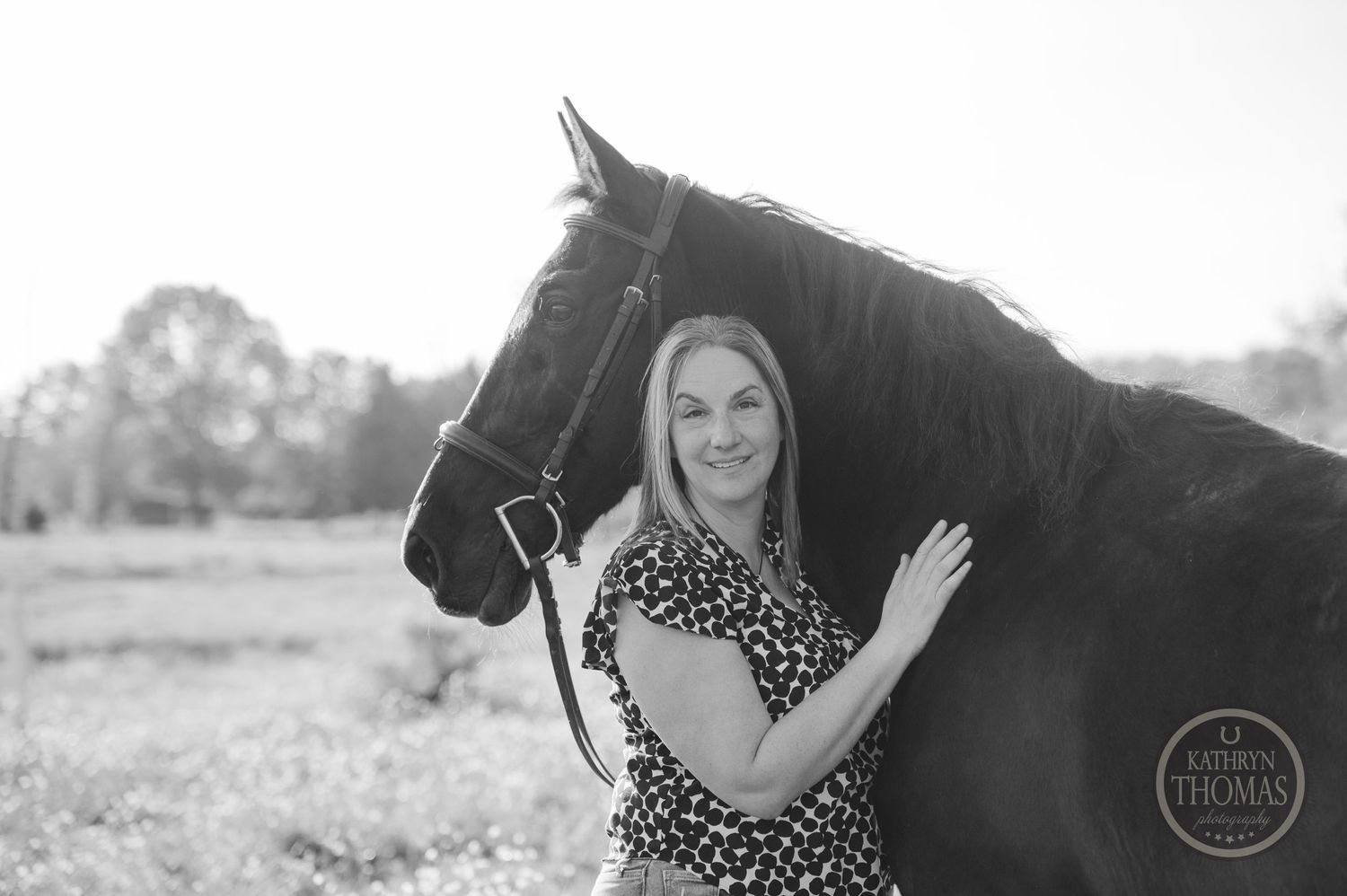 Black and white portrait of a woman posing with her majestic dark horse in a country field.