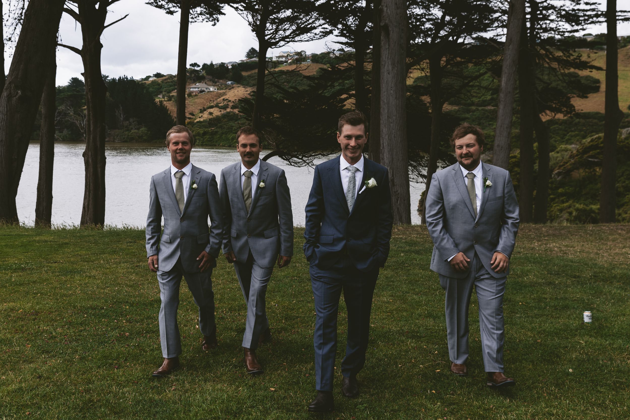 Groomsmen in matching gray suits walking by a lakeside with pine trees.