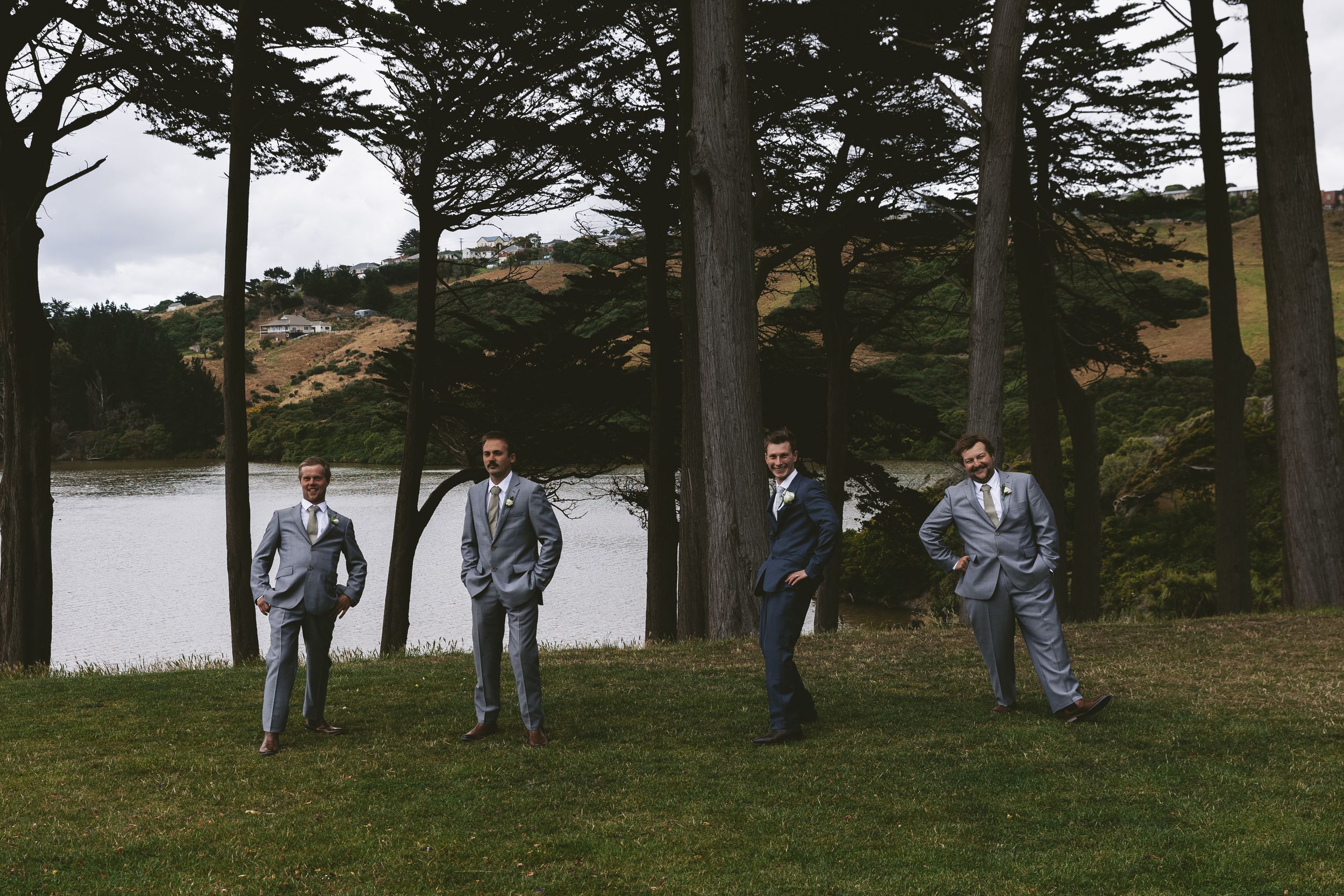 Wedding party in gray suits posing casually by a lake with pine trees.