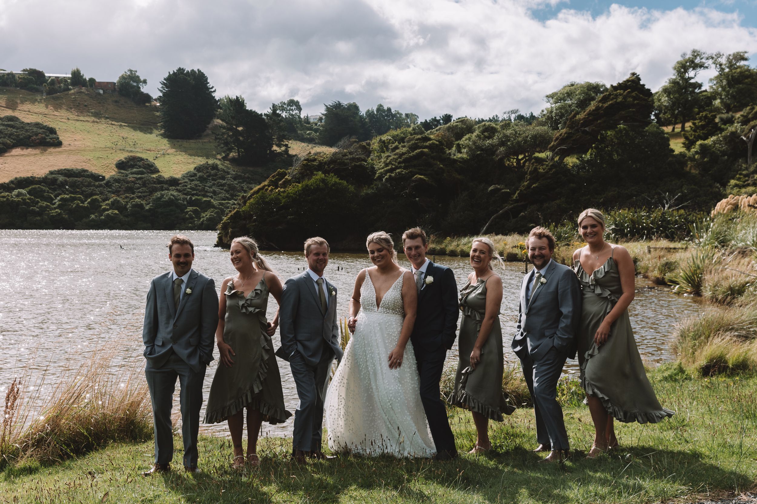 Wedding party lined up for a formal group photo beside a scenic lake.
