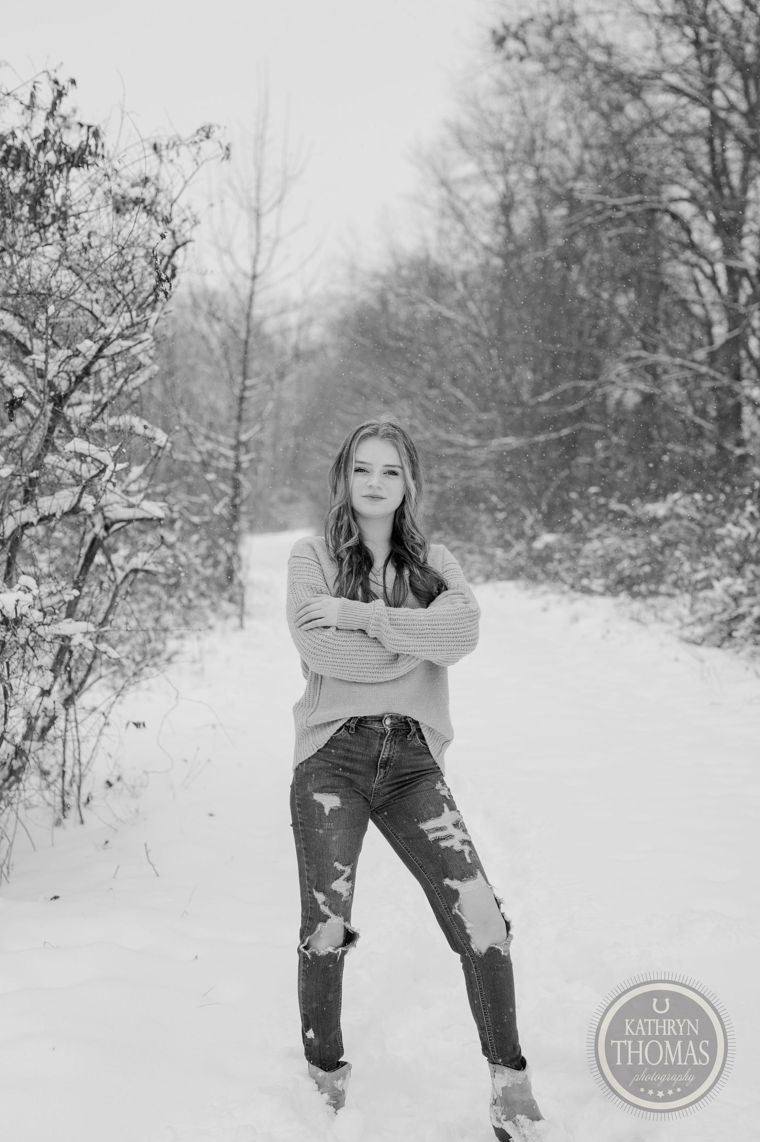 A black and white winter portrait of someone posing confidently in ripped jeans on a snowy path.
