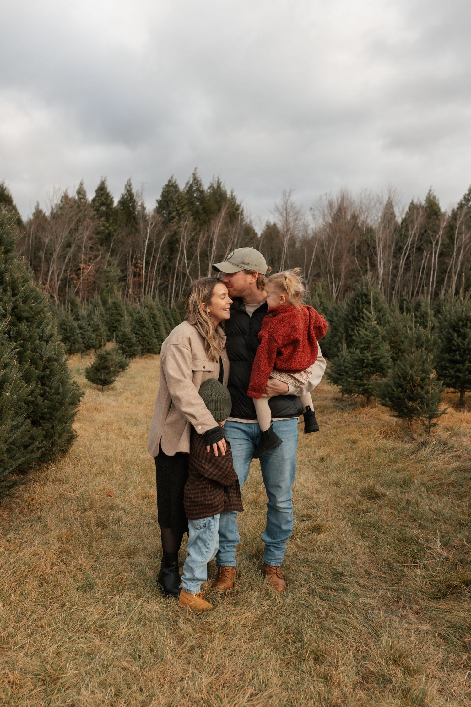 A couple shares a kiss while holding their toddler in a red sweater at a Christmas tree farm on a cloudy day.