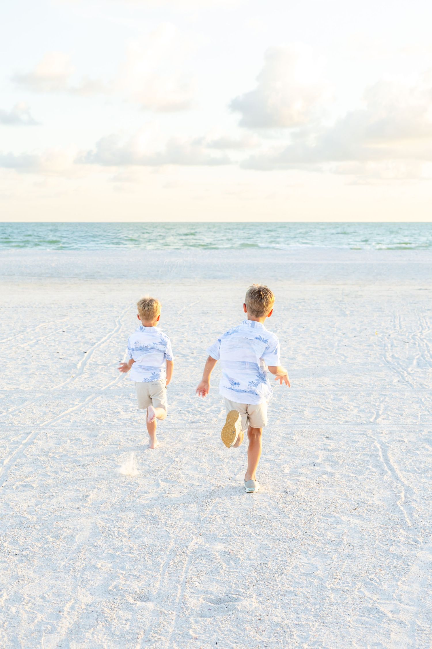 Two young boys in white shirts run barefoot toward the ocean on a white sandy beach at sunset.