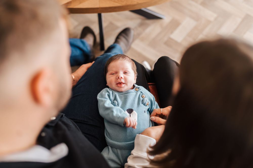 A smiling baby in a light blue sweater sits on a dark couch while adults interact nearby.