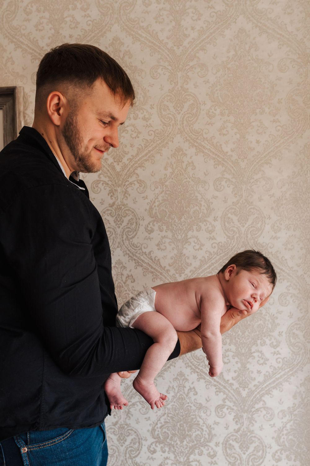 A person in a black shirt holds a sleeping newborn baby against a wallpapered background.