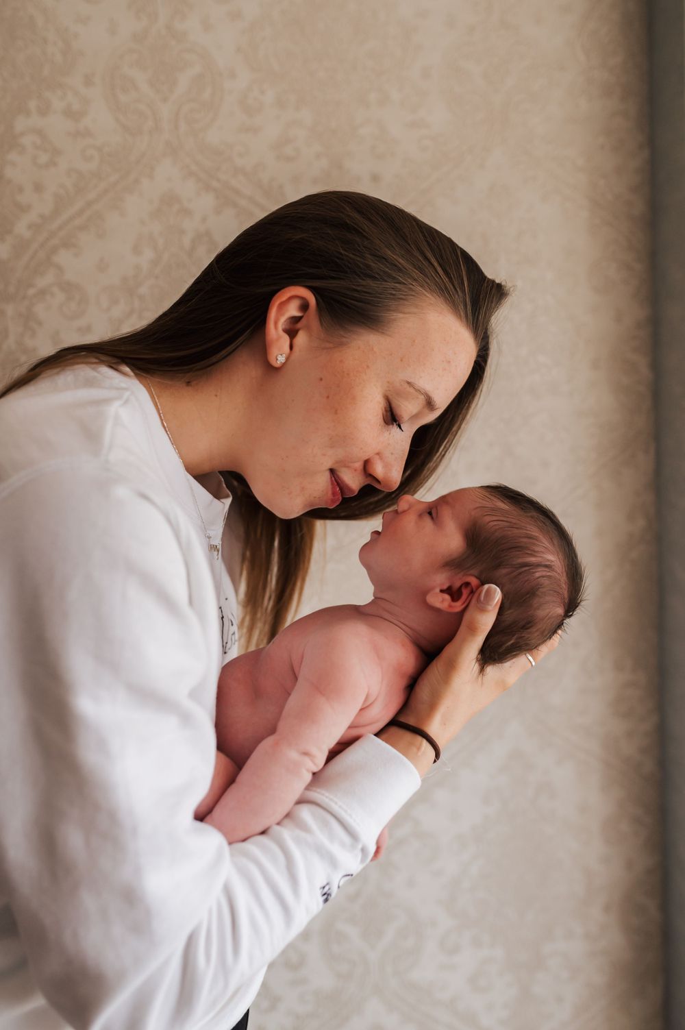 A tender moment captured between a mother in a white sweater and her newborn baby against a textured beige background.