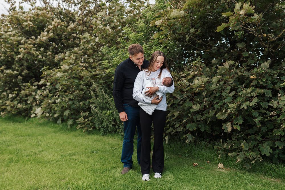 A couple stands on grass near a hedge while holding a baby in a white outfit on a sunny day.