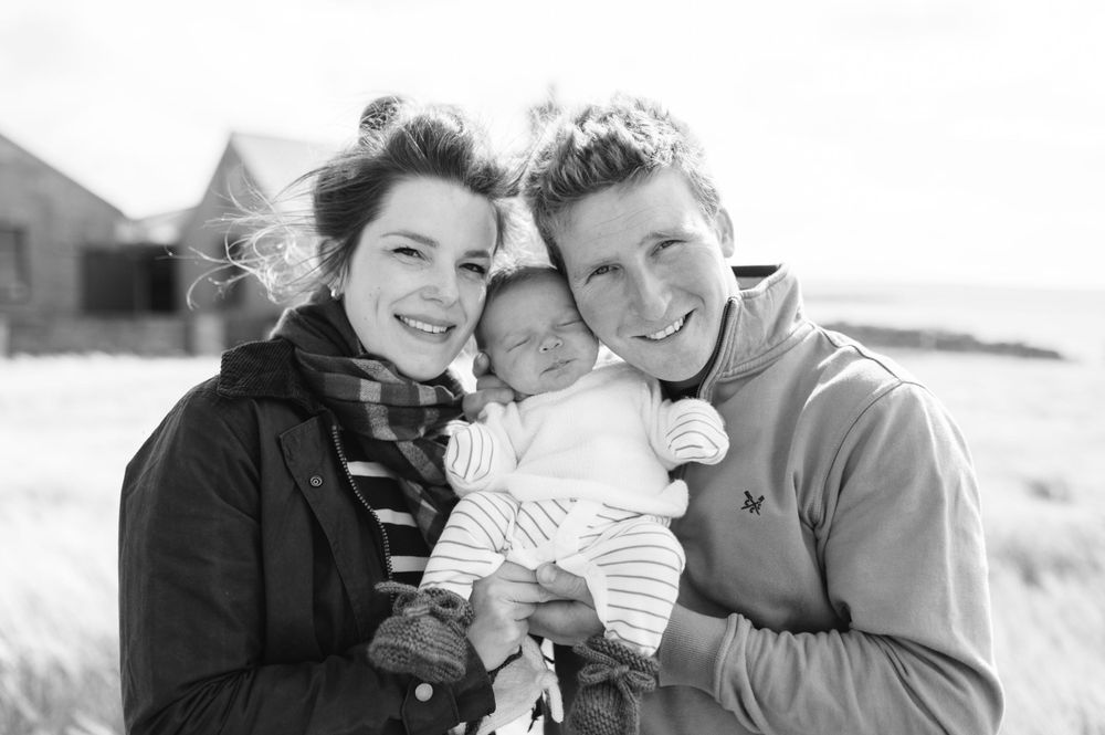 A black and white portrait of two adults holding a baby between them while smiling outdoors on a sunny day.