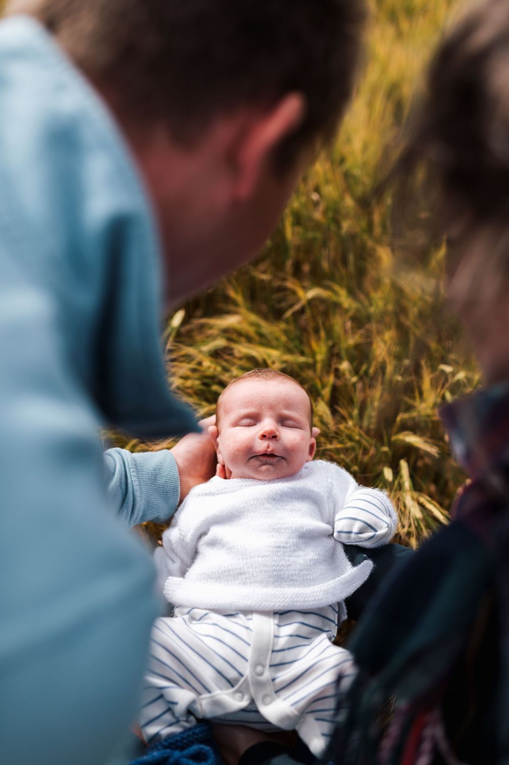 Baby in white clothing lies on grass while being photographed by adults in blue clothing on a sunny day.