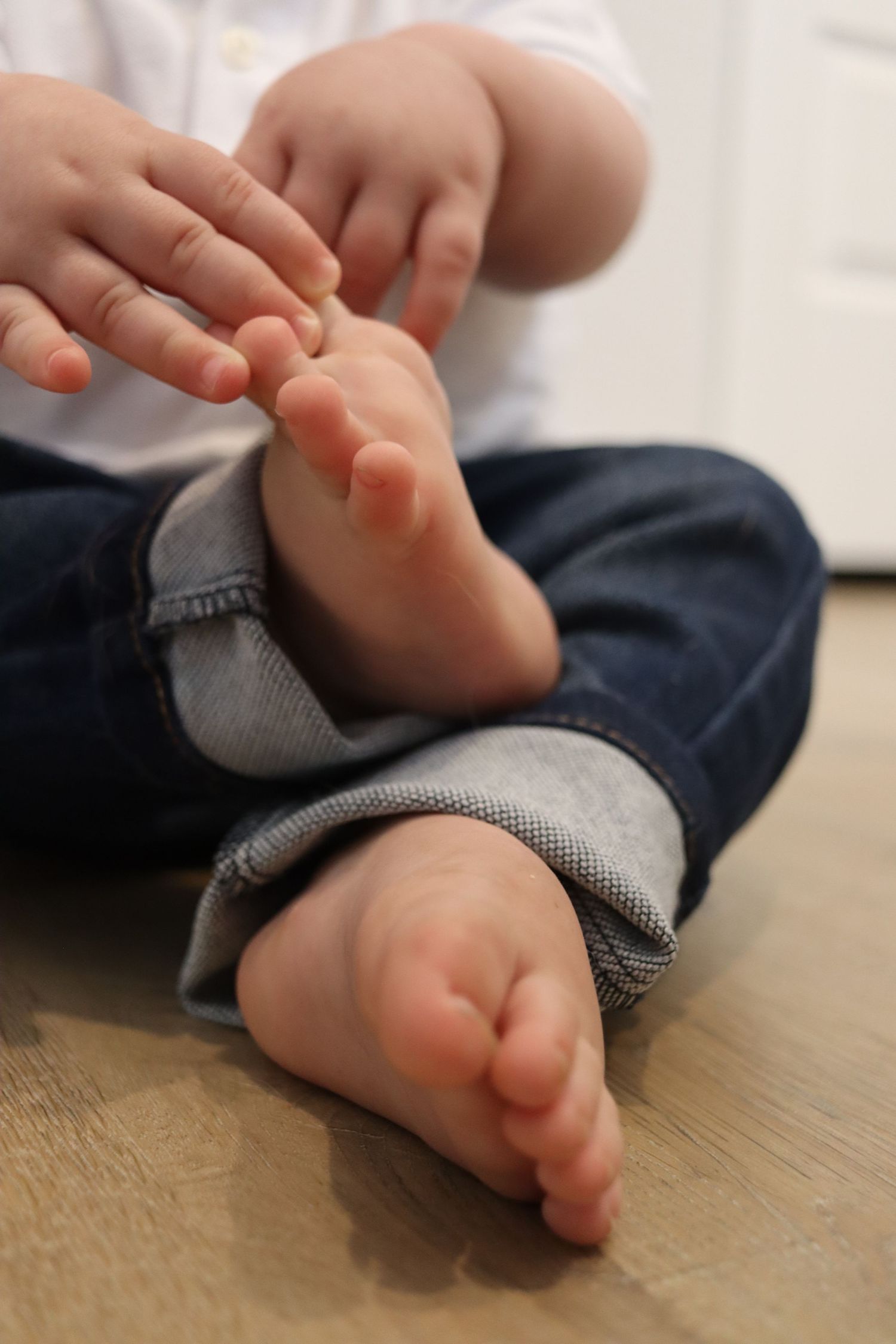 Close-up of baby feet and hands sitting cross-legged on hardwood floor in jeans.