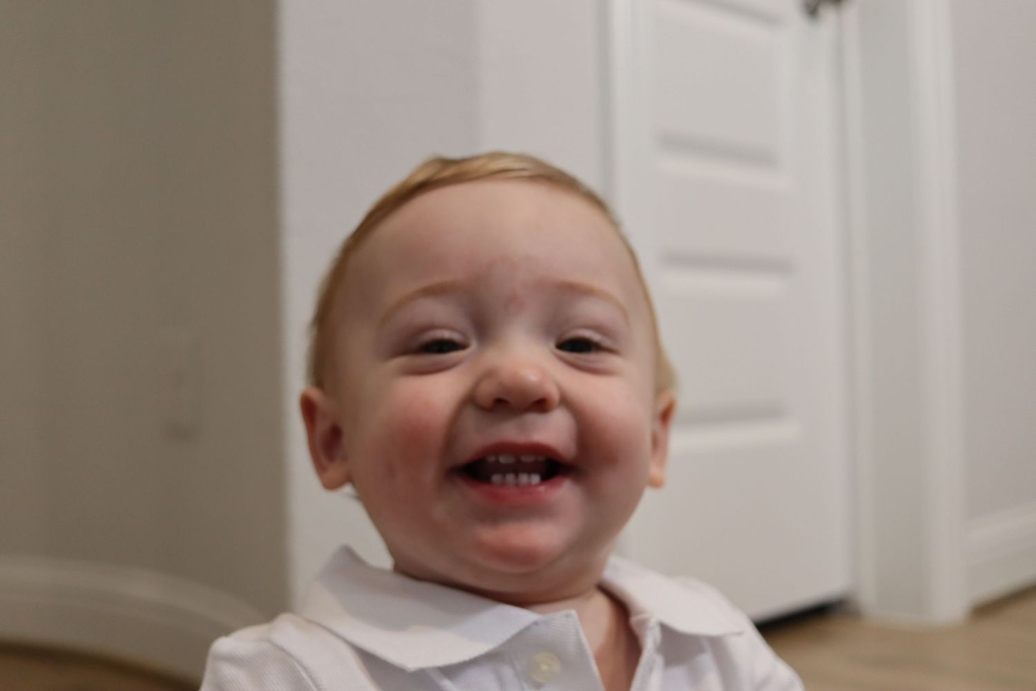 Adorable smiling baby with blonde hair wearing a white shirt indoors.