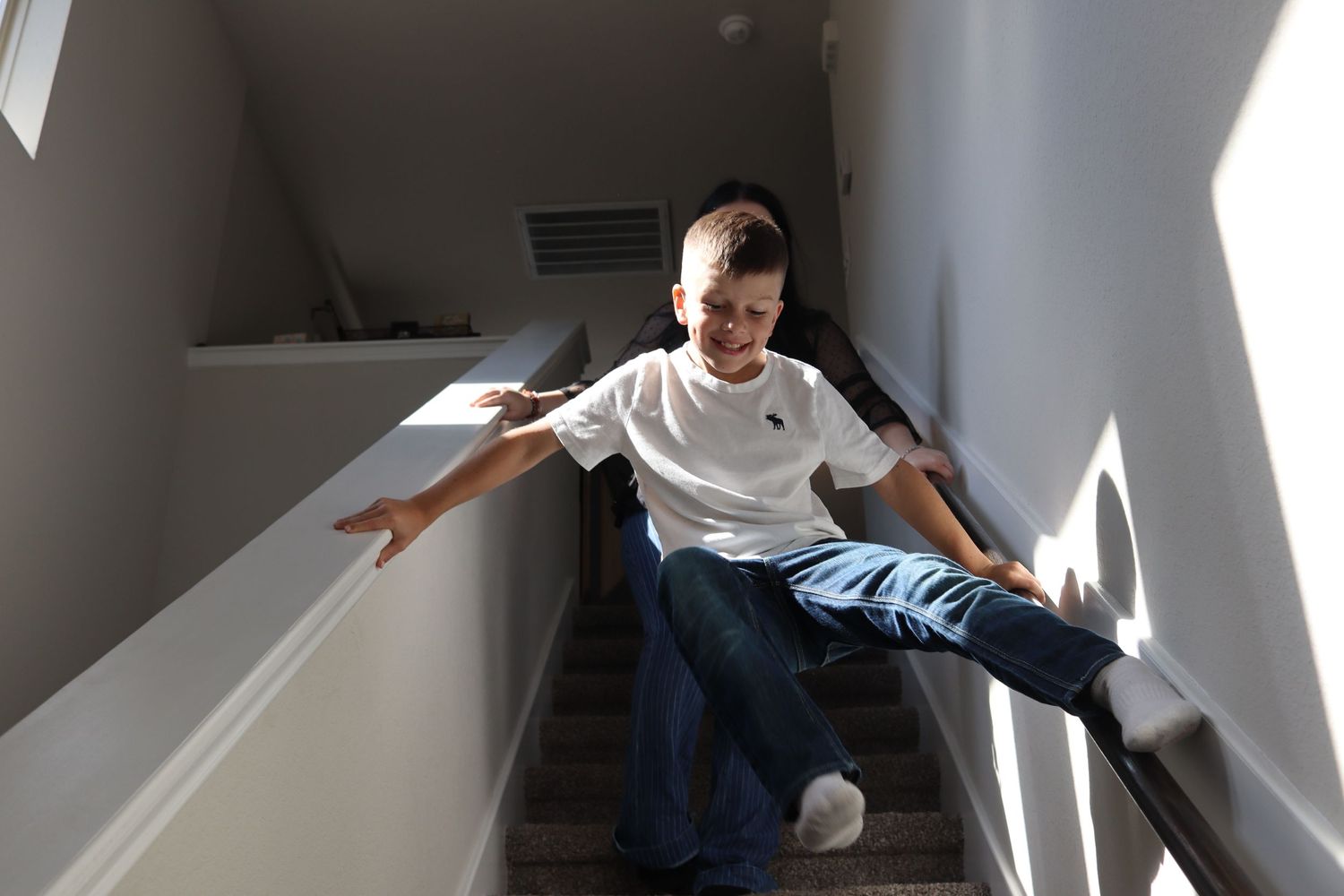 Smiling boy sliding down indoor staircase railing with arms outstretched in sunlit home.