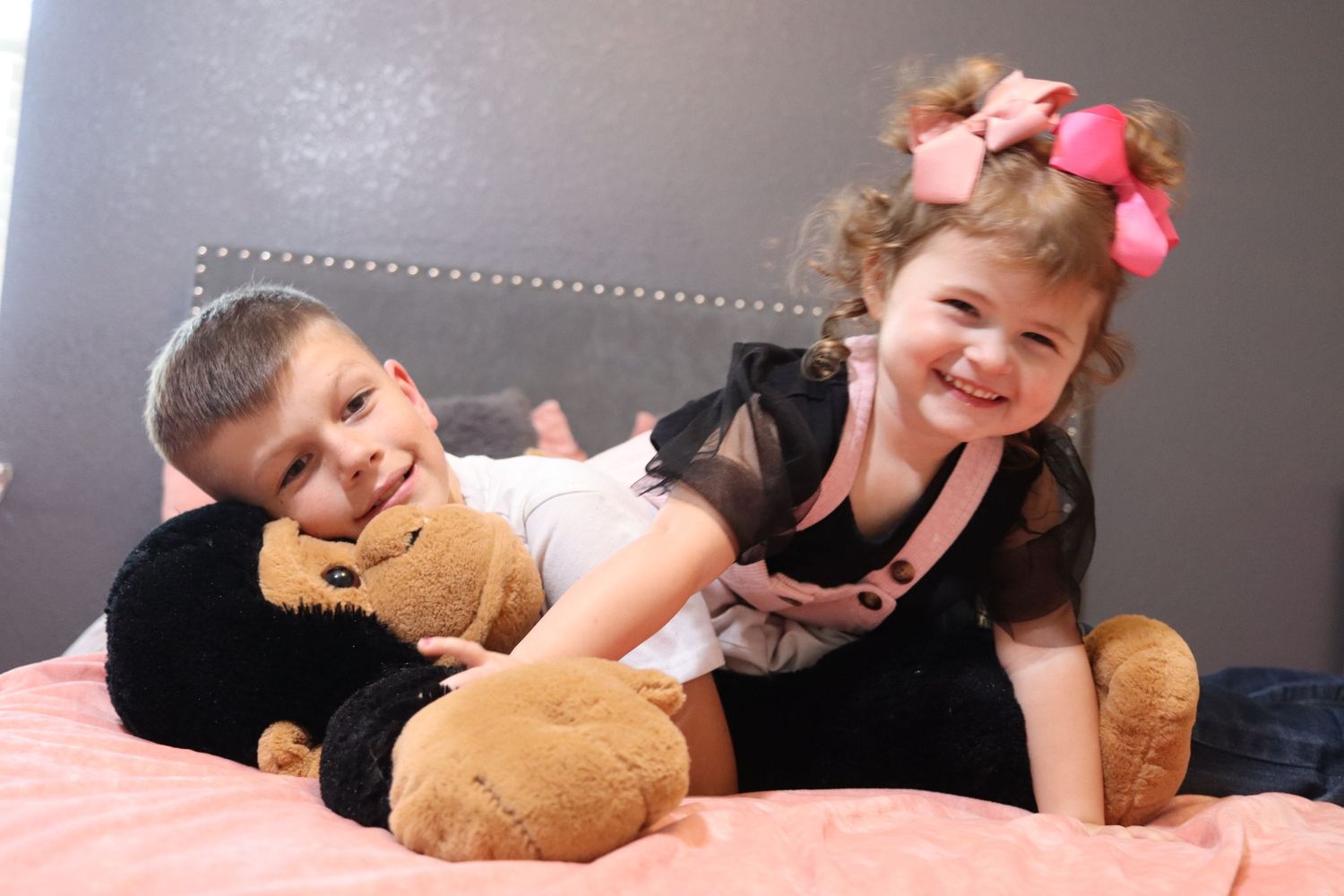 Two young children laughing and playing with a stuffed teddy bear on a pink bed.