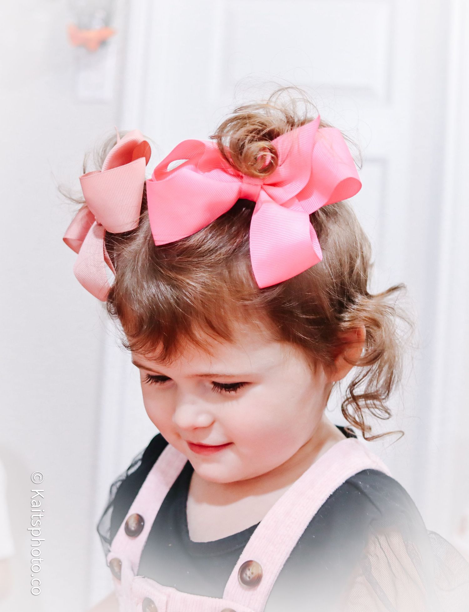 An adorable toddler girl with curly hair wears large pink ribbon bows in a soft portrait photo.