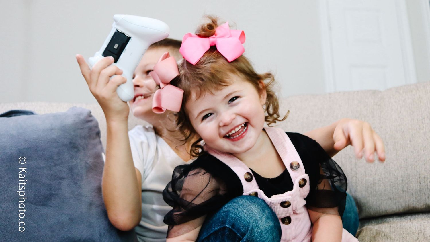 Two young girls laugh joyfully on a couch, one holding a phone for a selfie, wearing pink hair bows.
