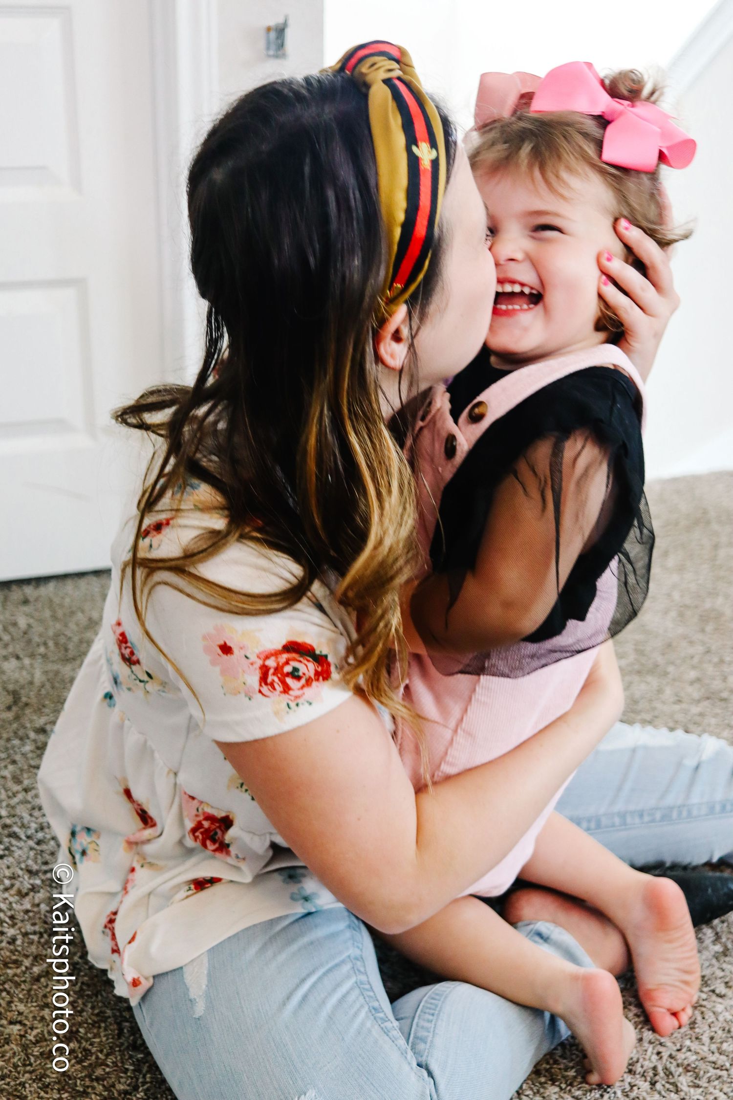 Mother kissing laughing toddler girl wearing pink bow on cheek indoors at home.