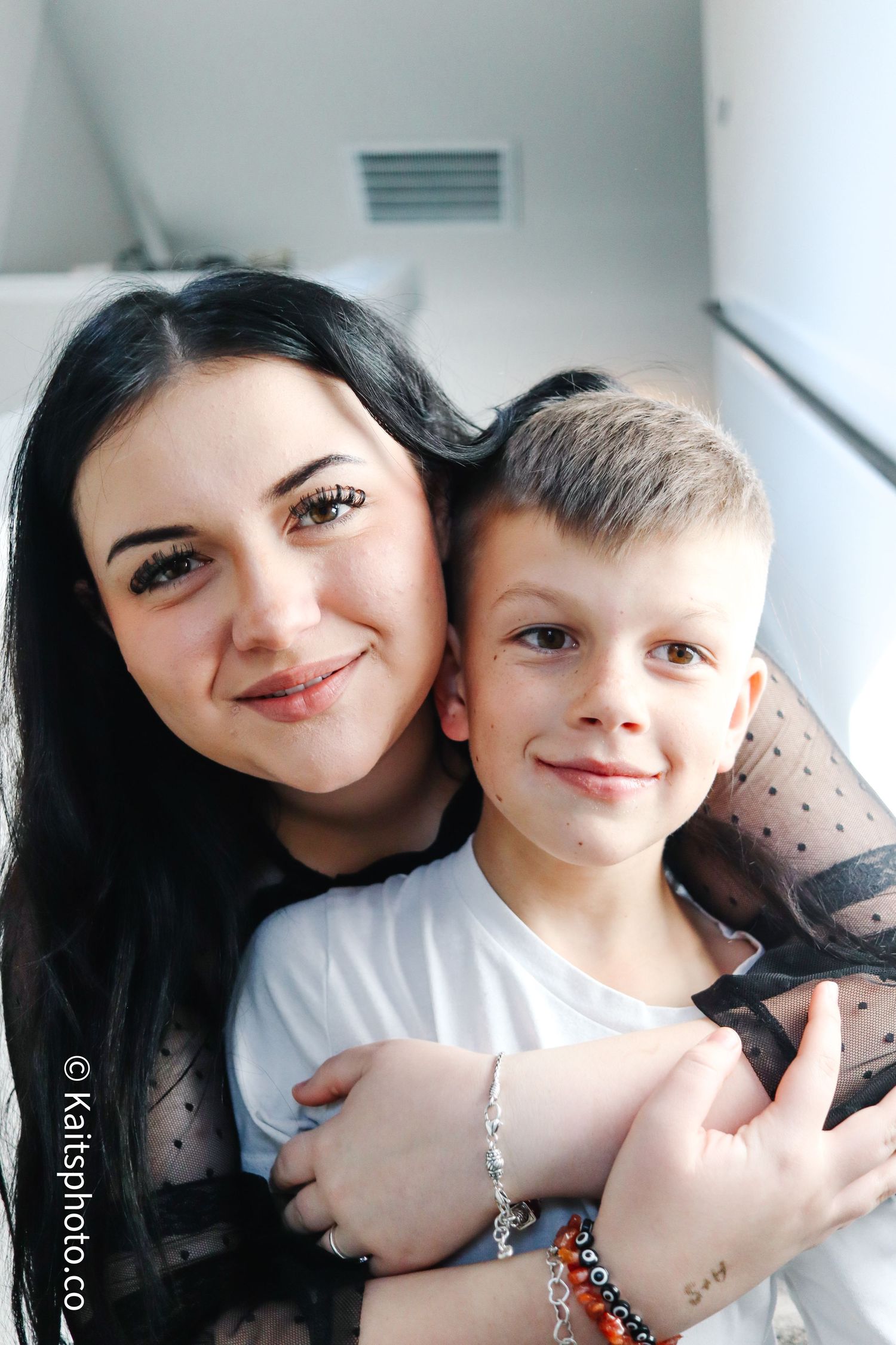 A young woman with dark hair smiles warmly while hugging a young boy in a bright indoor setting.