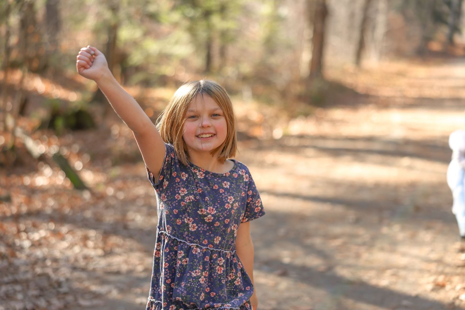 Happy blonde girl in floral dress raising fist on forest trail.