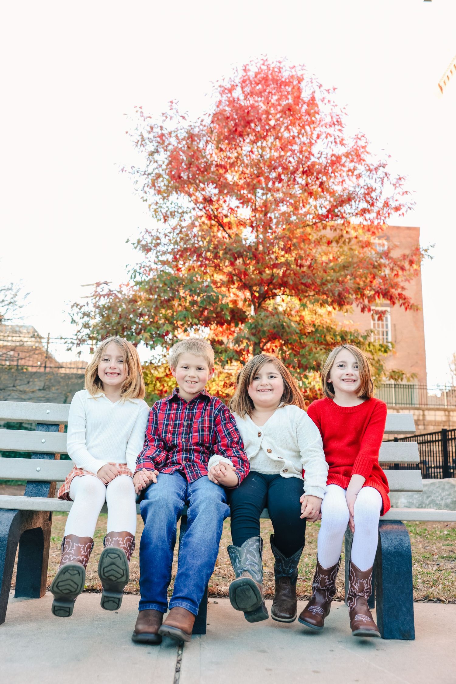Four children sitting on a bench in front of vibrant red autumn foliage wearing cowboy boots.