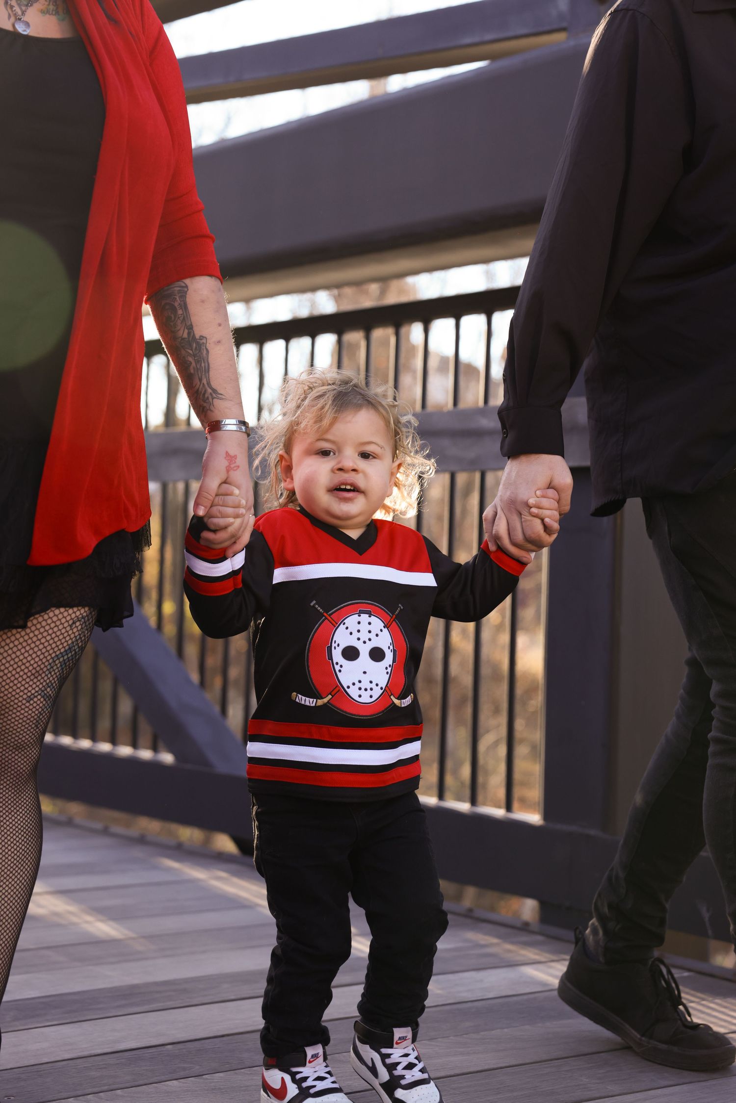 Toddler in hockey jersey walking holding parents hands on bridge.