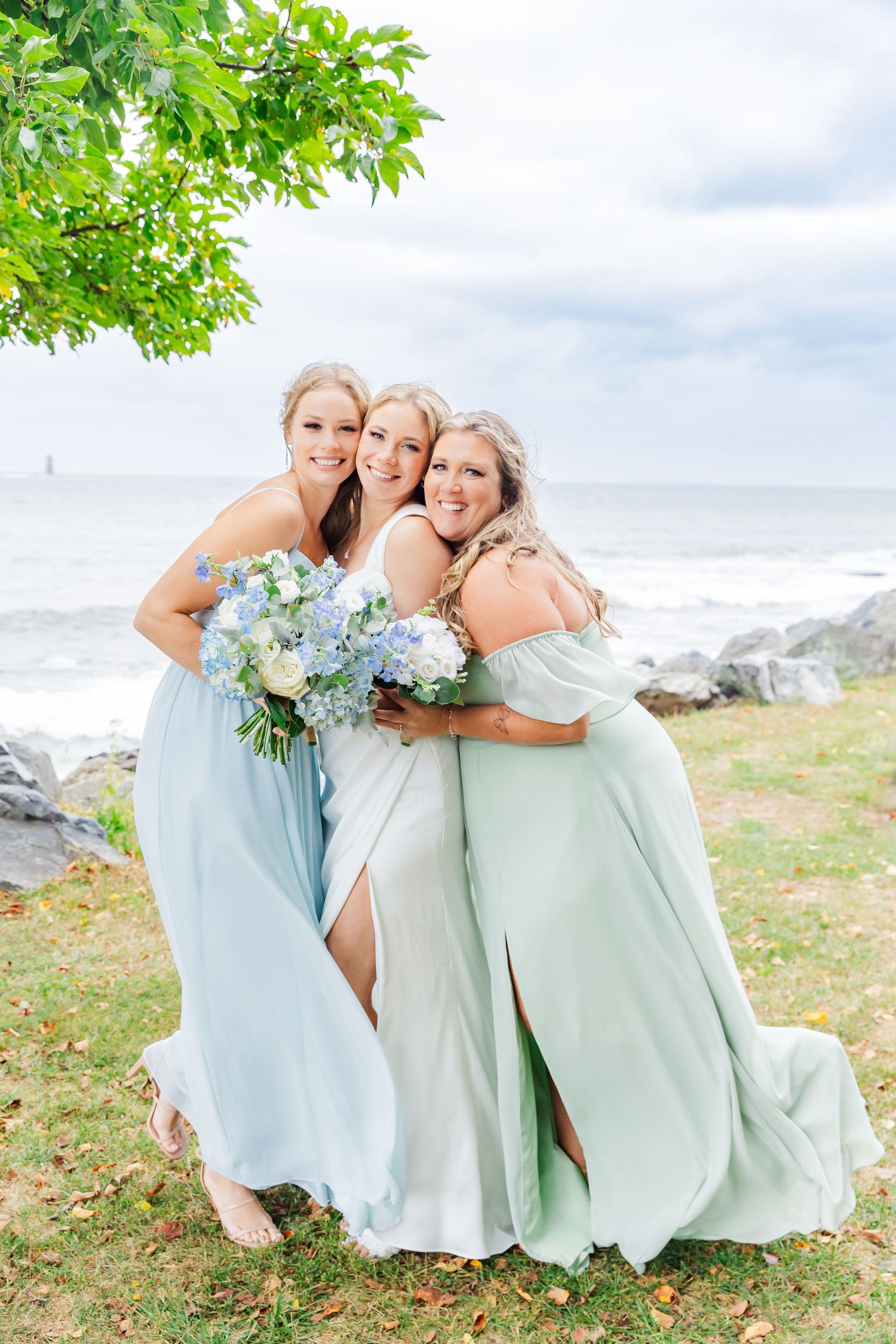 Three bridesmaids in light blue dresses pose with bouquets by the ocean.