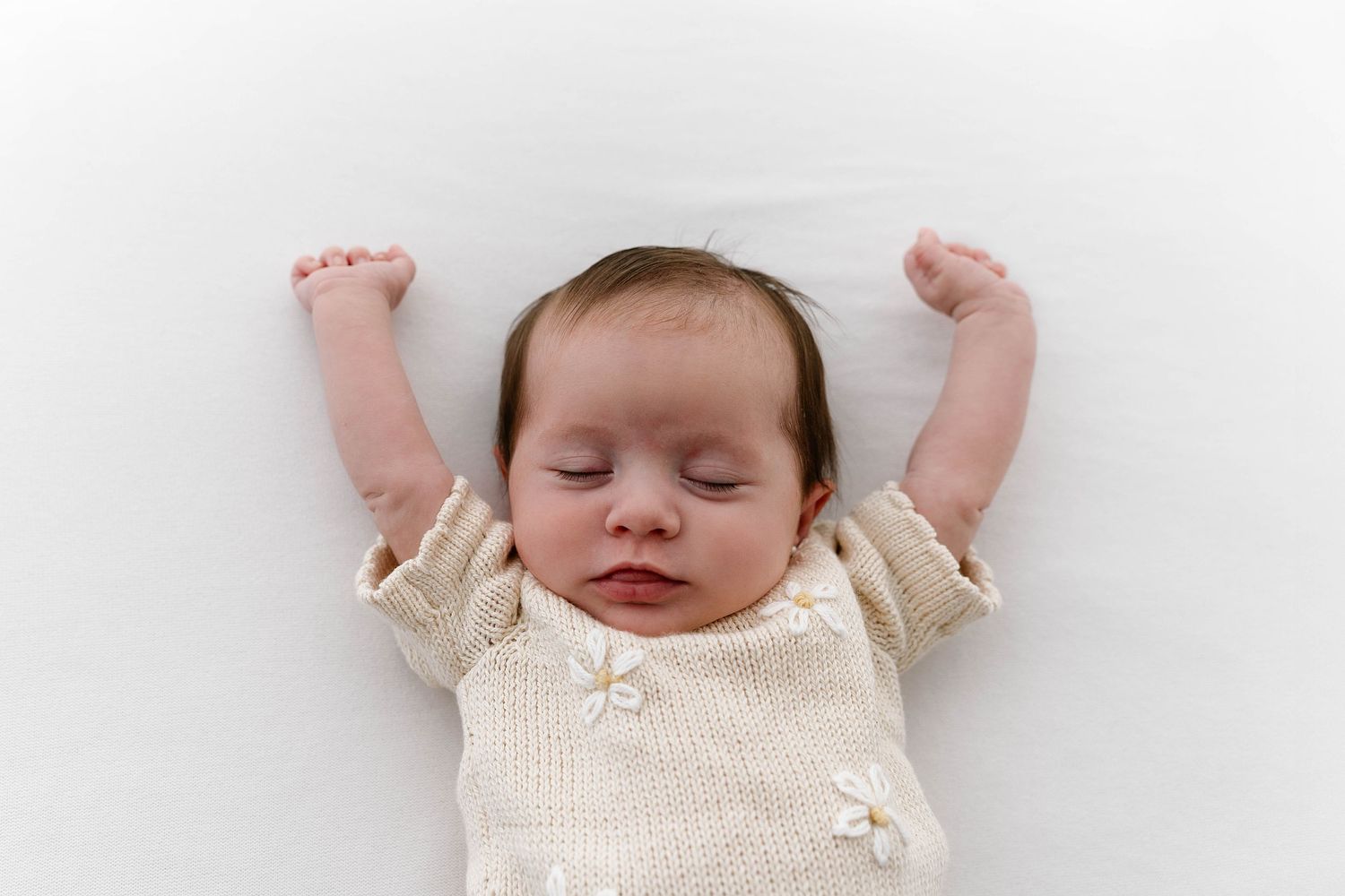 Sleeping baby in cream outfit stretches arms up against white background.