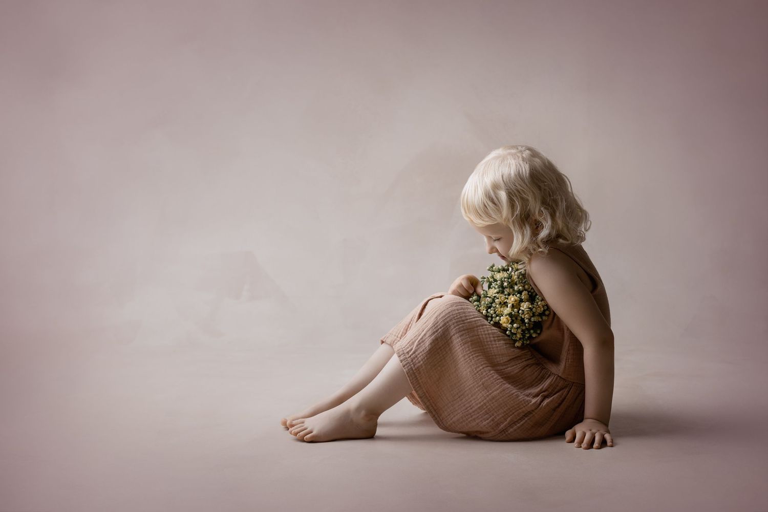 Child wearing a tan summery top sitting barefoot on a neutral pink backdrop in a studio setting.