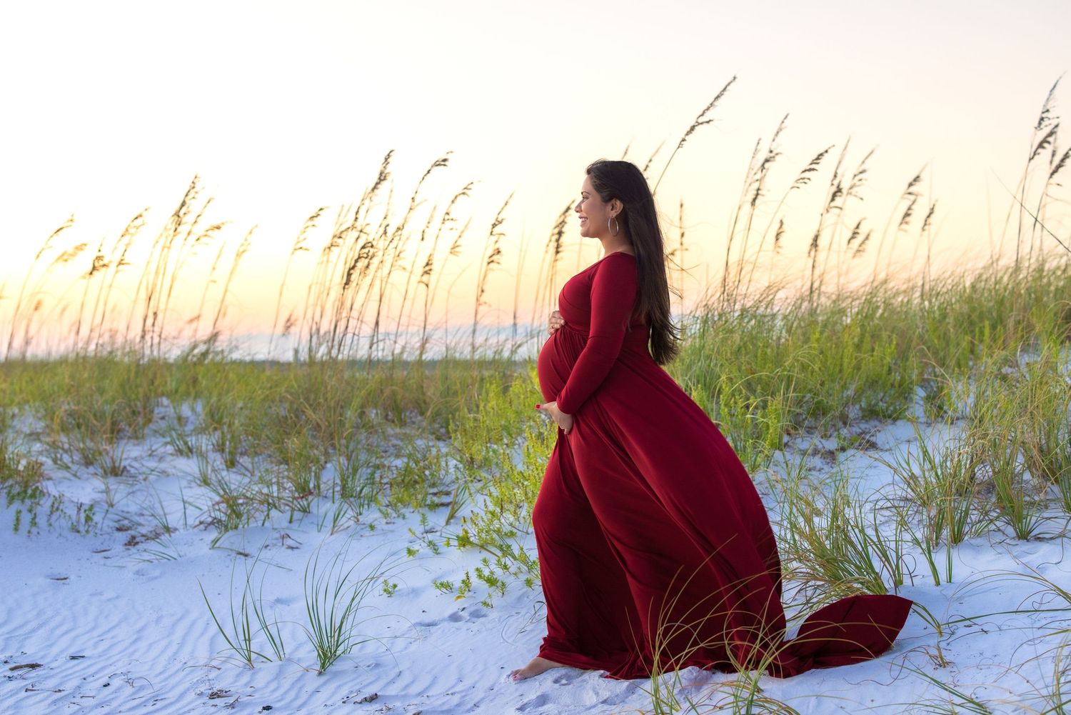 Elegant figure in flowing red velvet dress stands gracefully against beach dunes at sunset.