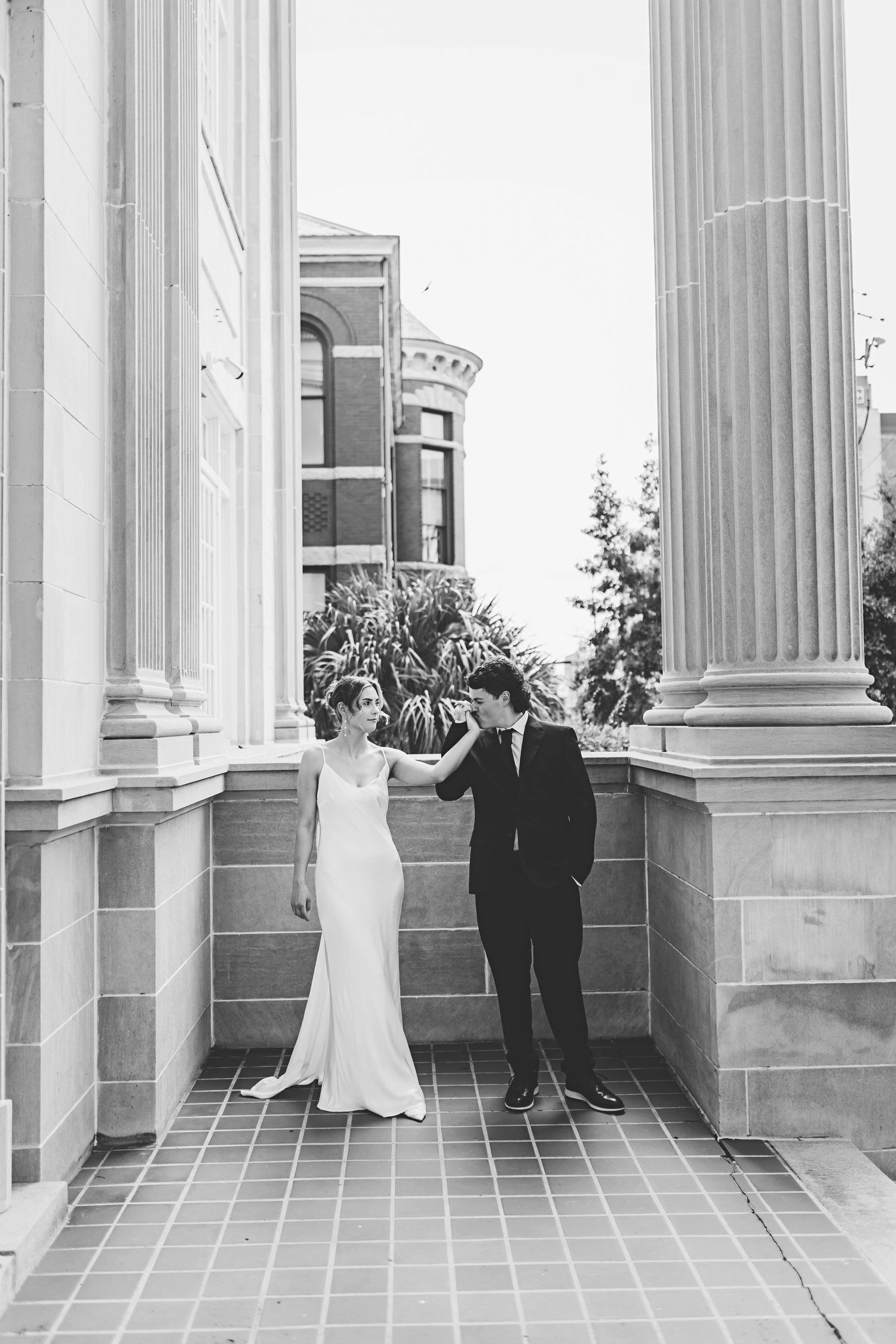 A couple in wedding attire stand between classical white columns on a tiled terrace of an elegant building.