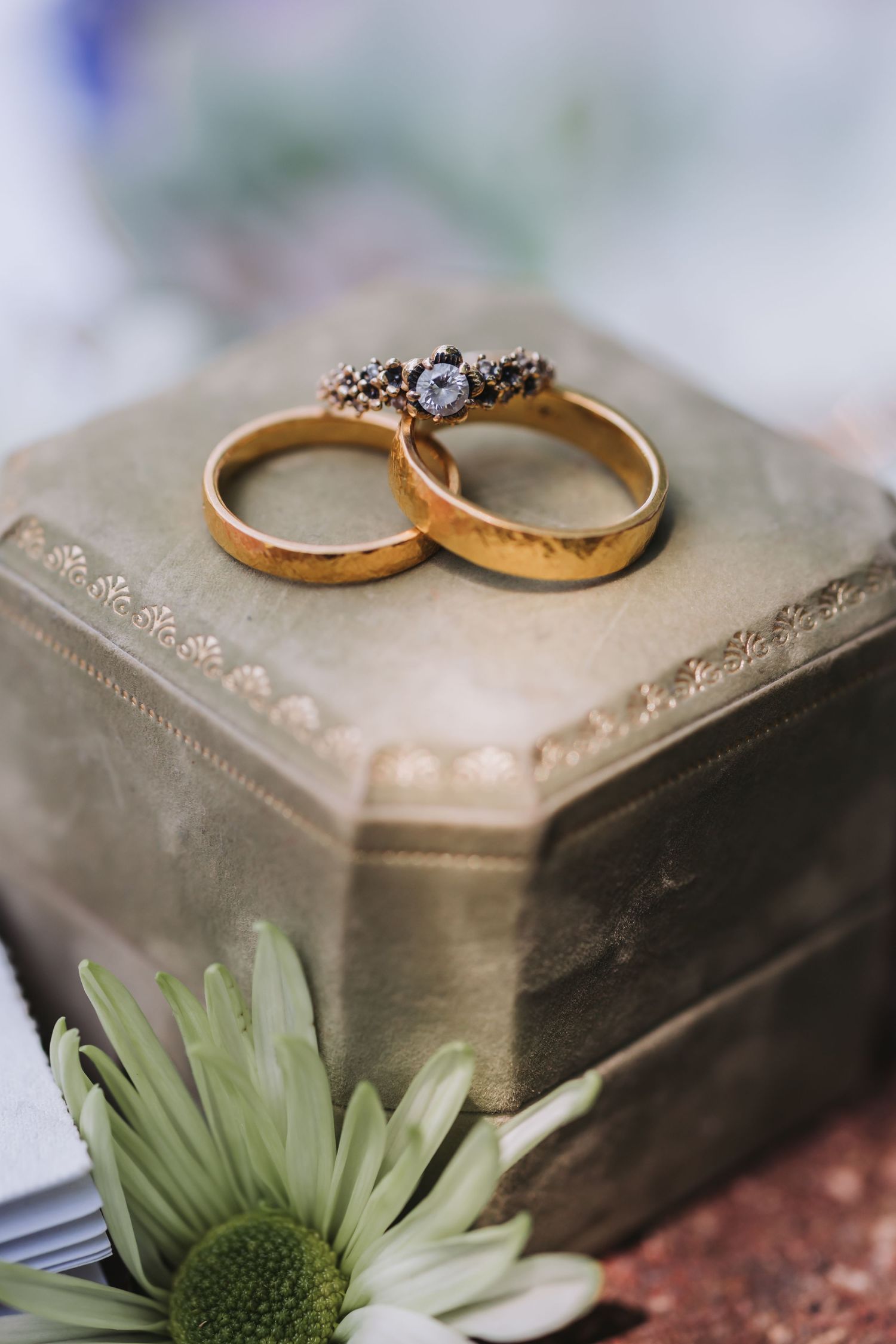Wedding rings with a vintage-style engagement ring displayed on a pewter jewelry box with a green flower accent.
