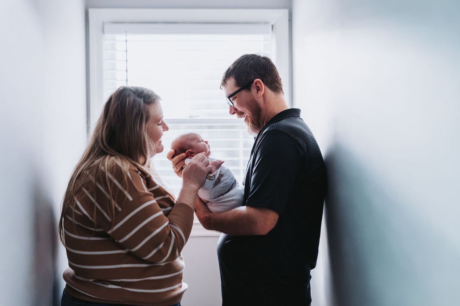 Two people stand near a window while holding and looking lovingly at a newborn baby wrapped in white.