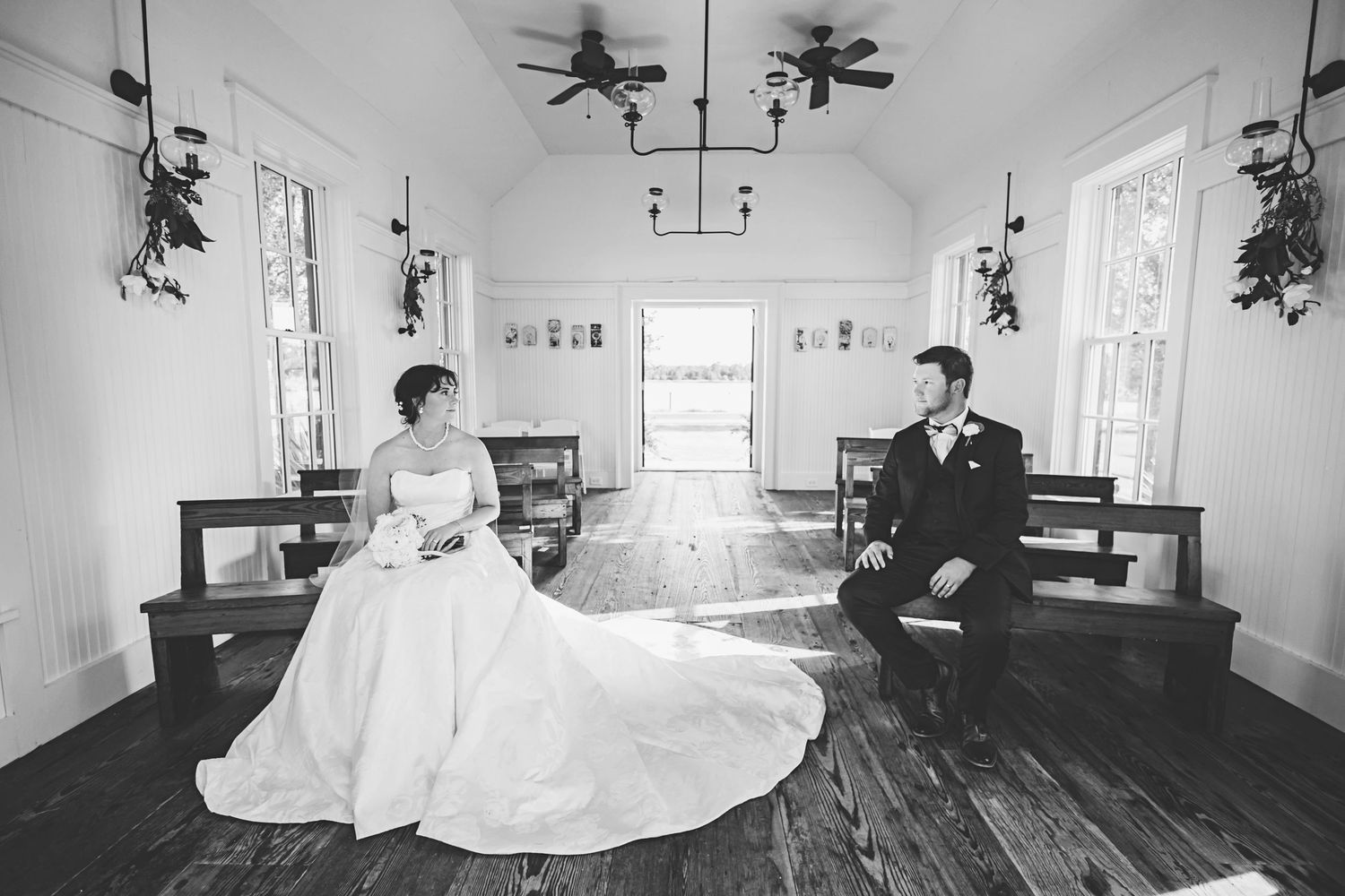 A vintage black and white photograph of a couple seated apart in a rustic chapel with wooden floors and white walls.