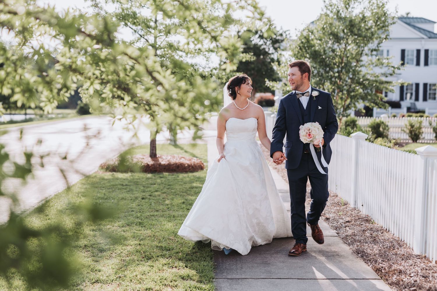 A couple in formal wedding attire walks together along a tree-lined garden path on a sunny afternoon.