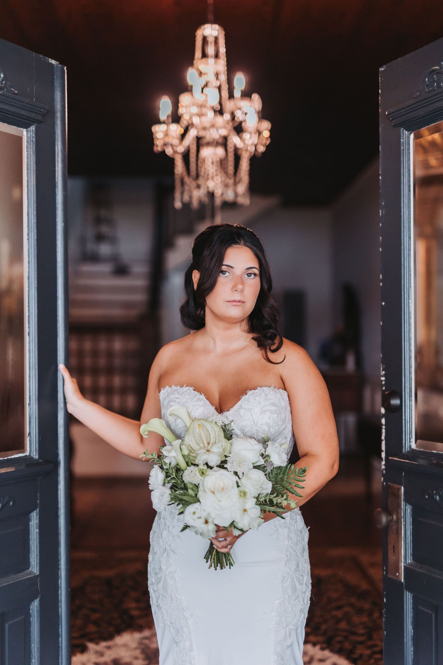 A bride in a white lace wedding dress holds a bouquet of white flowers against an elegant dark interior with chandeliers.