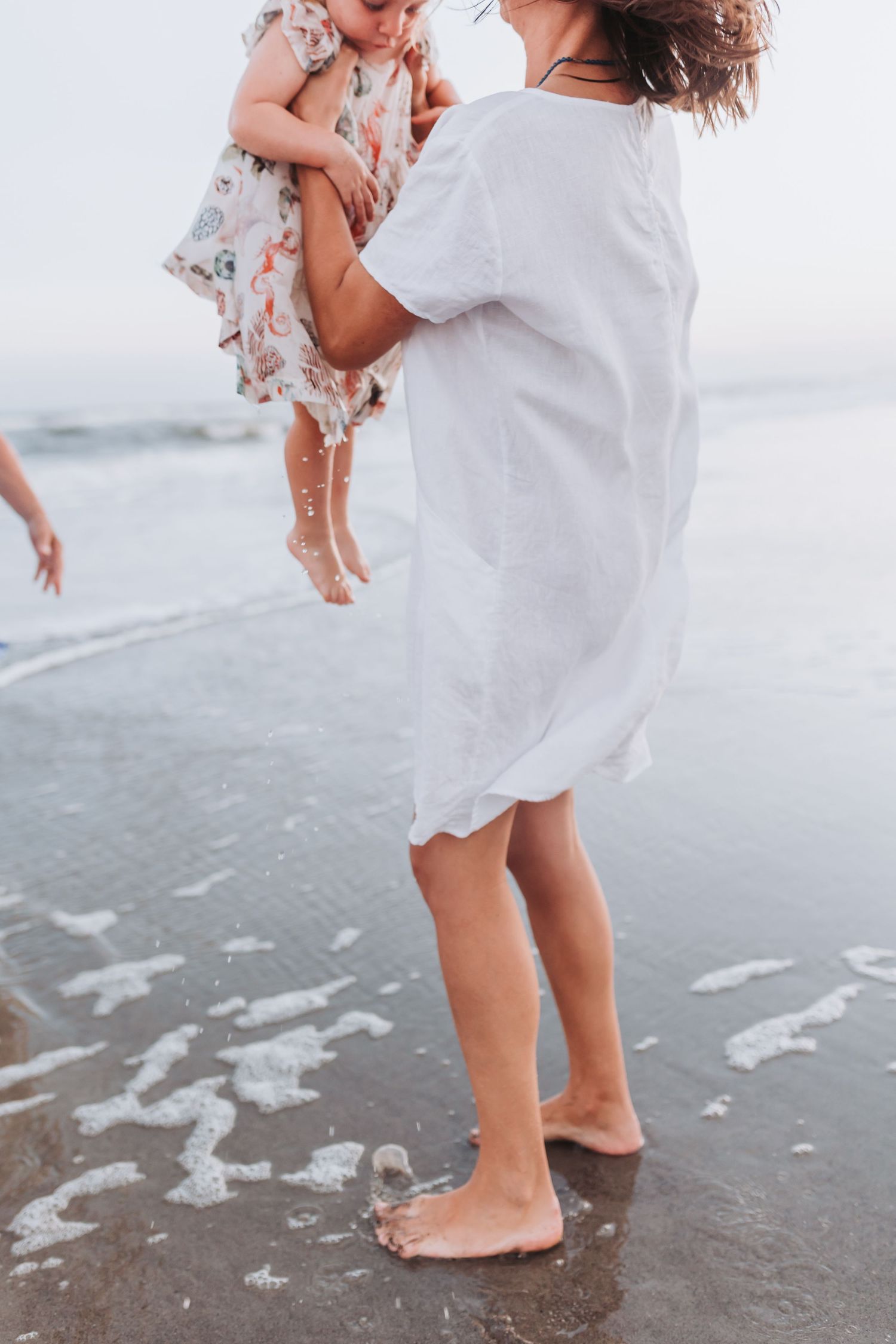 A person in a white dress lifts a small child while standing barefoot on a beach with gentle waves rolling in.