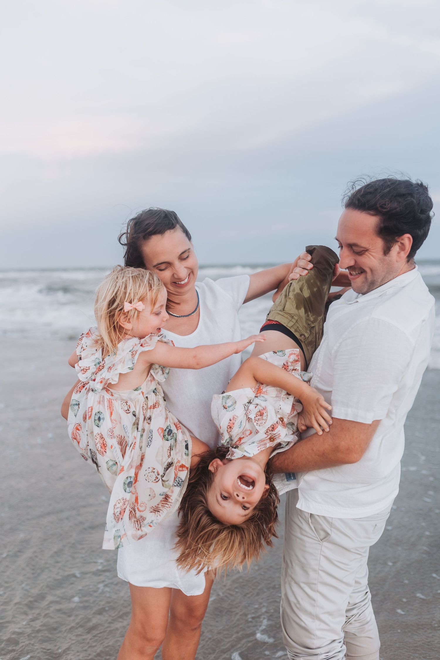 A family enjoys a playful moment together on the beach at sunset with children in floral dresses.