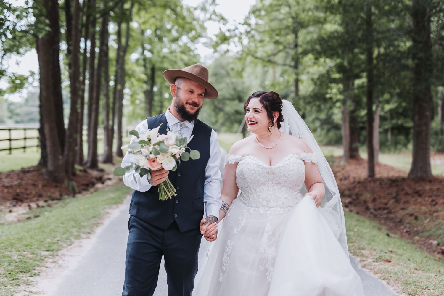 A joyful couple in wedding attire walks hand in hand down a tree-lined dirt path holding a white floral bouquet.