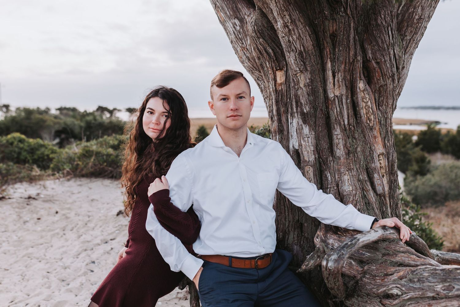 A couple in burgundy dress and white shirt embrace under a large tree during a romantic outdoor photoshoot.
