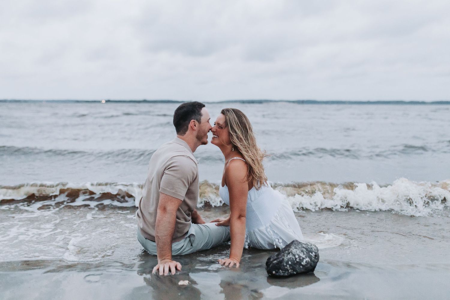 A romantic couple sits in shallow ocean water as waves crash behind them during a beach photoshoot.