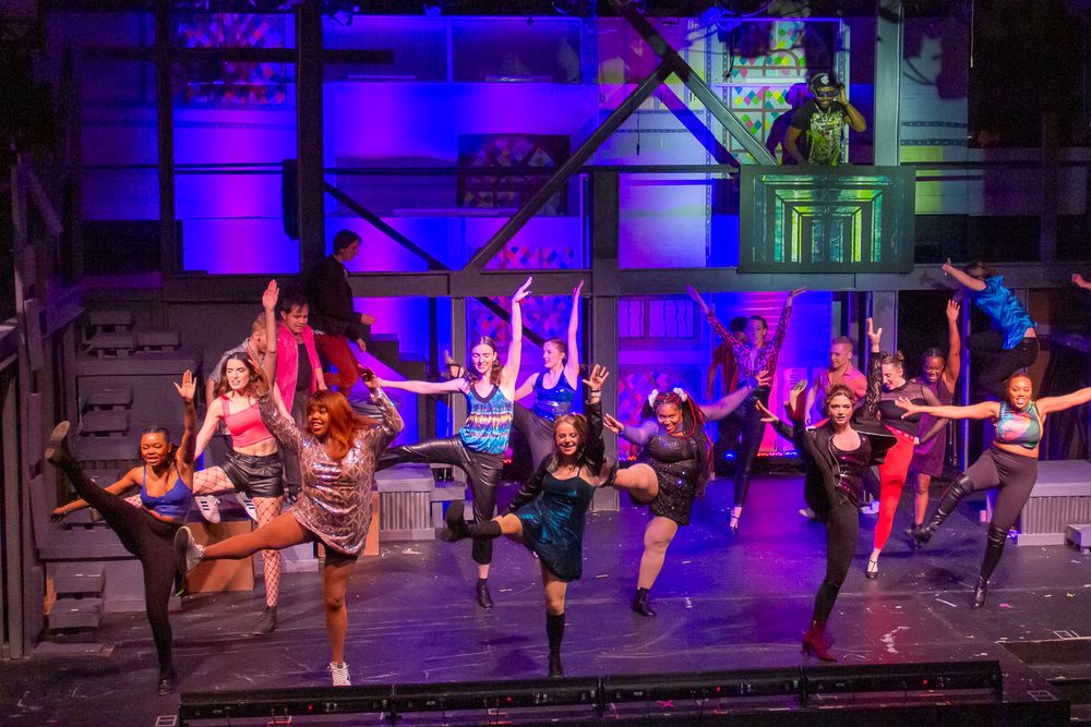 Dancers perform a high-energy ensemble number on stage with dramatic purple lighting.